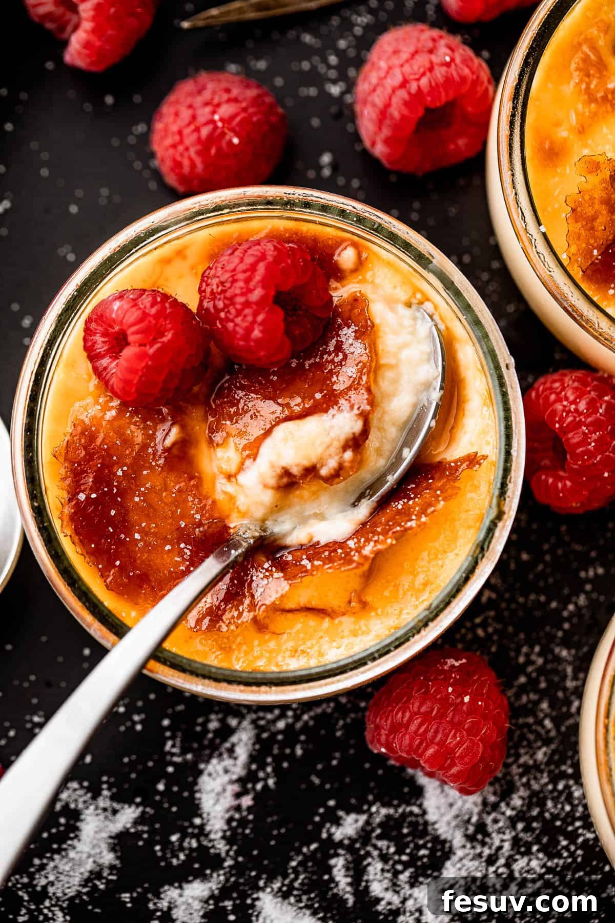 Foolproof Creme Brulee 2 overhead shot of creme brulee in a glass jar, topped with two raspberries, and a spoon inside the jar.