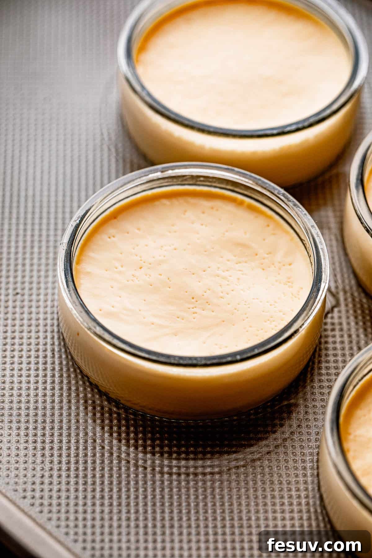 Side view of two creme brulee filled ramekins in a baking pan with water, ready for the oven.