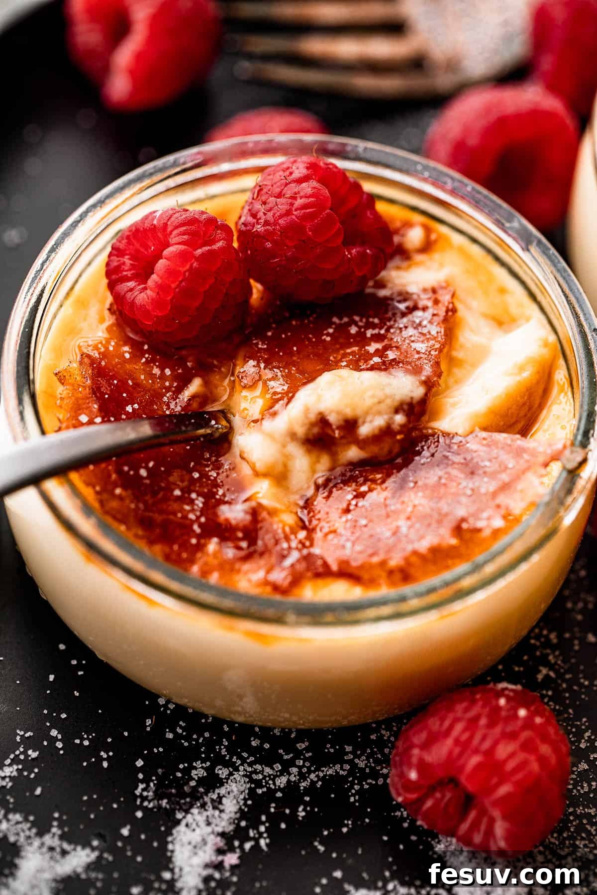 Side shot of a single creme brulee in a glass jar, garnished with two fresh raspberries, and a small spoon resting inside.
