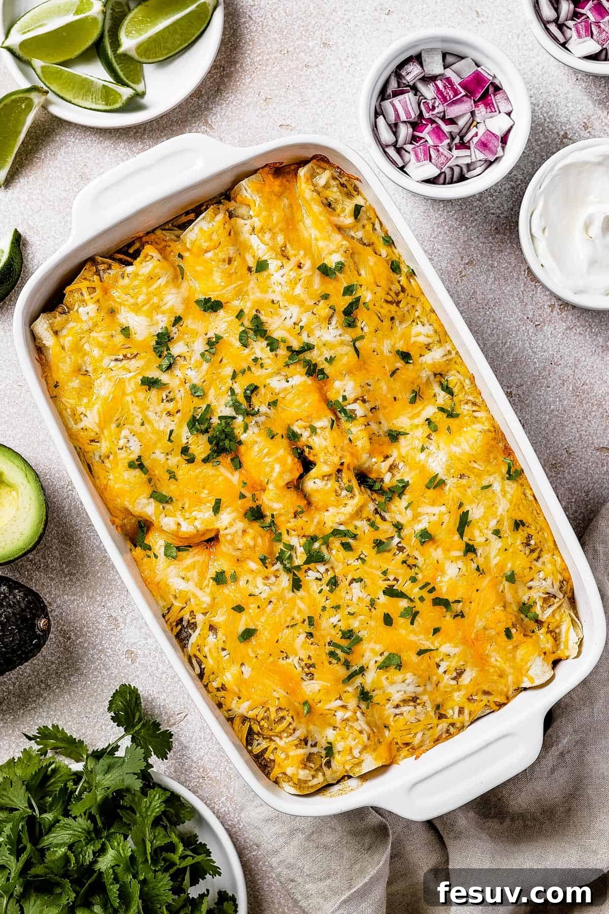 Smothered Green Chili Chicken Enchiladas 2 Overhead shot of perfectly baked salsa verde chicken enchiladas in a white baking dish, garnished with fresh cilantro, alongside small bowls of vibrant red onions, creamy sour cream, and zesty lime wedges for serving.