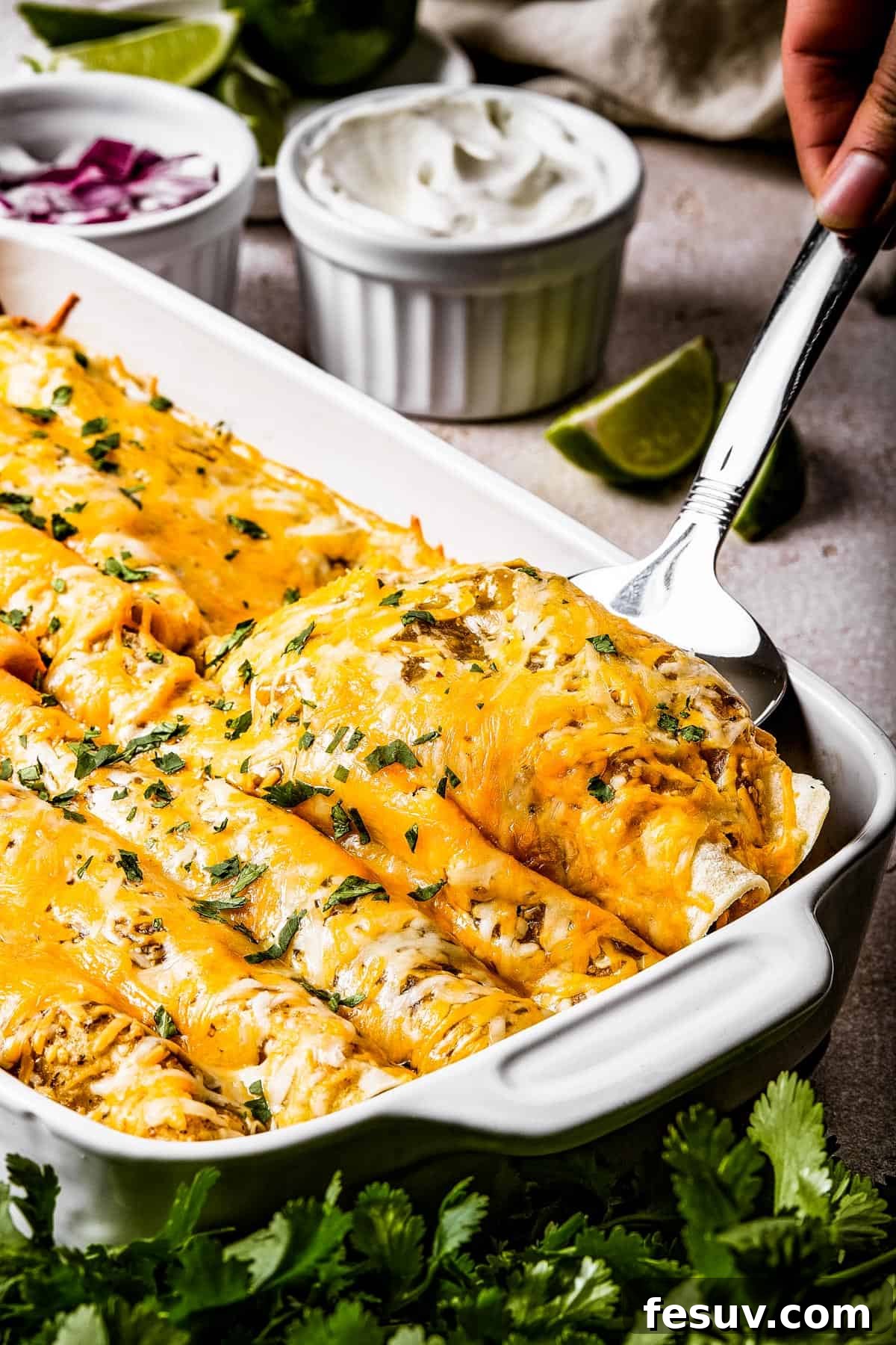 Smothered Green Chili Chicken Enchiladas 3 A glistening silver serving spoon lifts two perfectly baked and saucy enchiladas from a white baking dish, showcasing the melted cheese and vibrant green salsa.