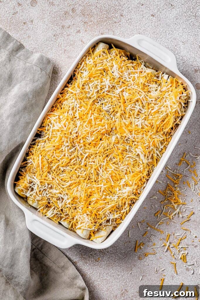 Smothered Green Chili Chicken Enchiladas 7 A pristine white baking dish filled with neatly rolled tortillas, generously topped with an even layer of shredded cheese, ready to be baked into golden perfection.