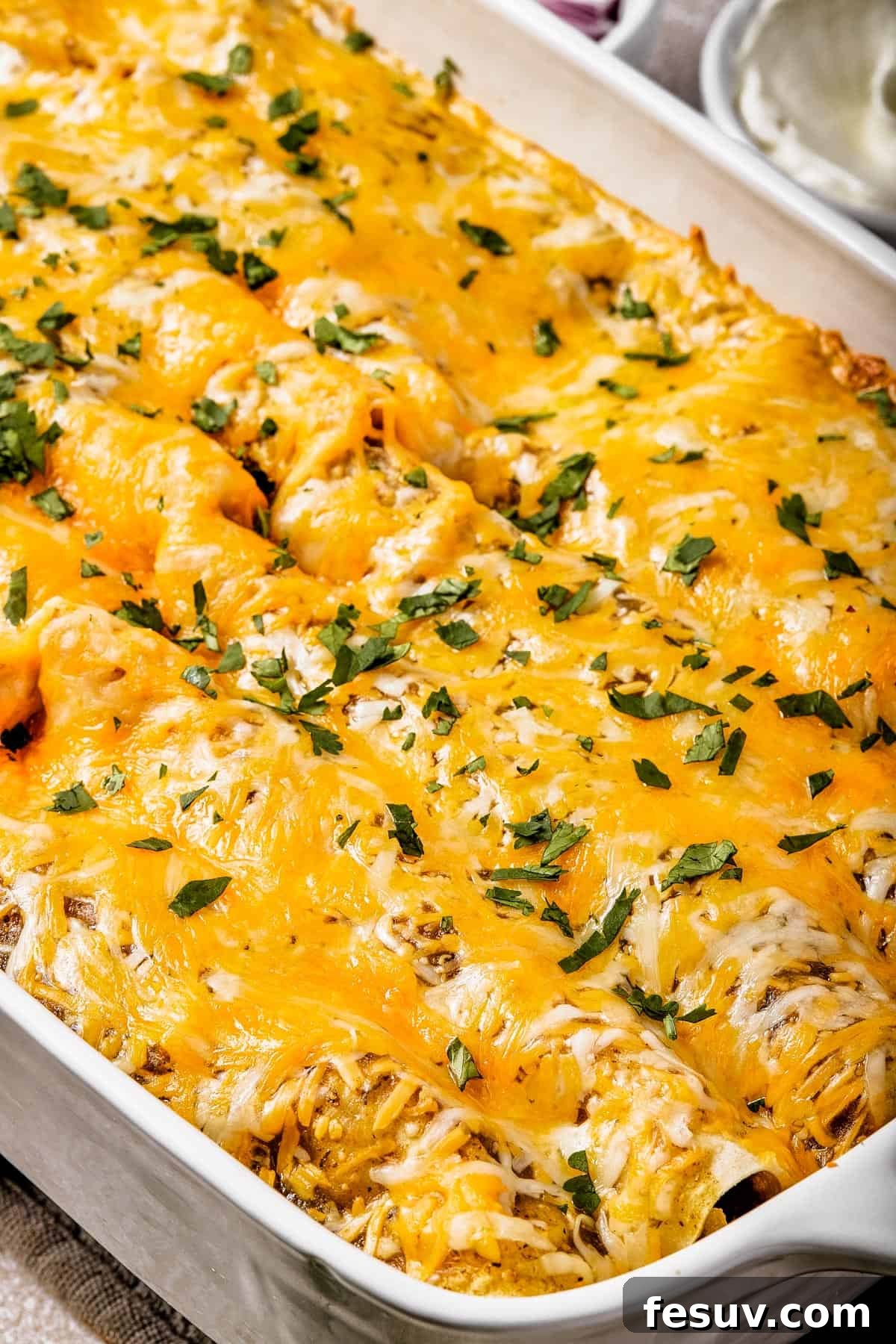 Smothered Green Chili Chicken Enchiladas 9 A tantalizing close-up shot of a baking dish filled with perfectly baked, golden-brown cheesy enchiladas, delicately sprinkled with fresh, vibrant green herbs for a pop of color and freshness.