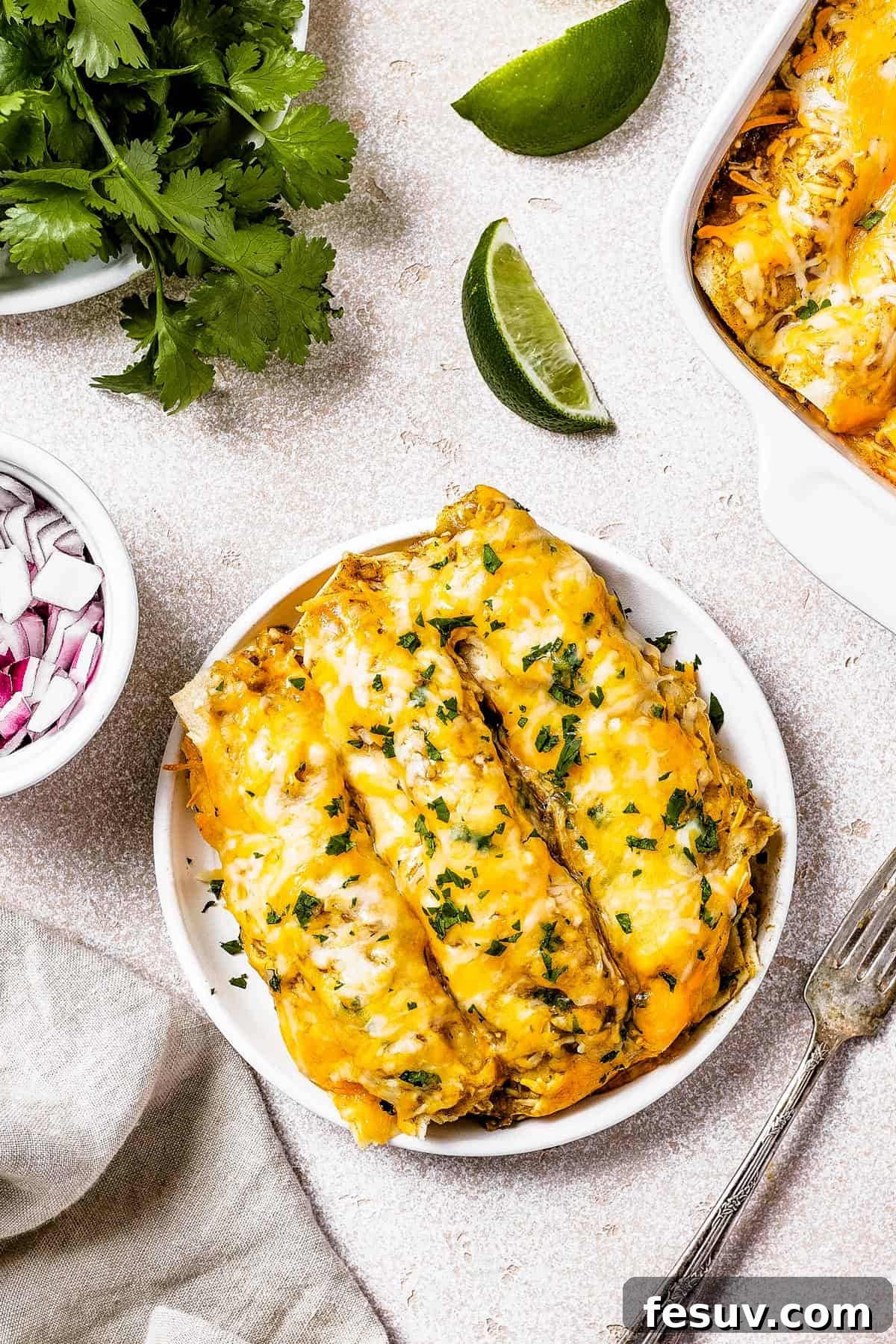 Smothered Green Chili Chicken Enchiladas 10 A single serving of golden-brown chicken enchiladas beautifully presented on a small white plate, artfully garnished with fresh herbs. It's perfectly positioned next to a rustic wooden dish of finely chopped red onion and a vibrant green bowl of fresh cilantro, ready for garnishing.