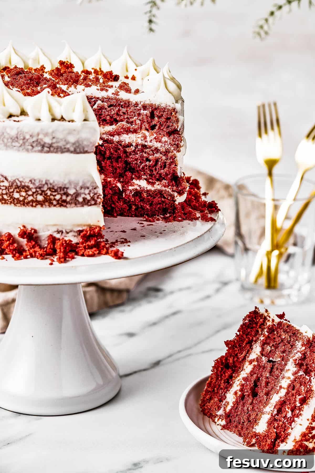 A slice of red velvet cake on a small plate next to the main cake on a stand, showcasing the moist interior and thick frosting.