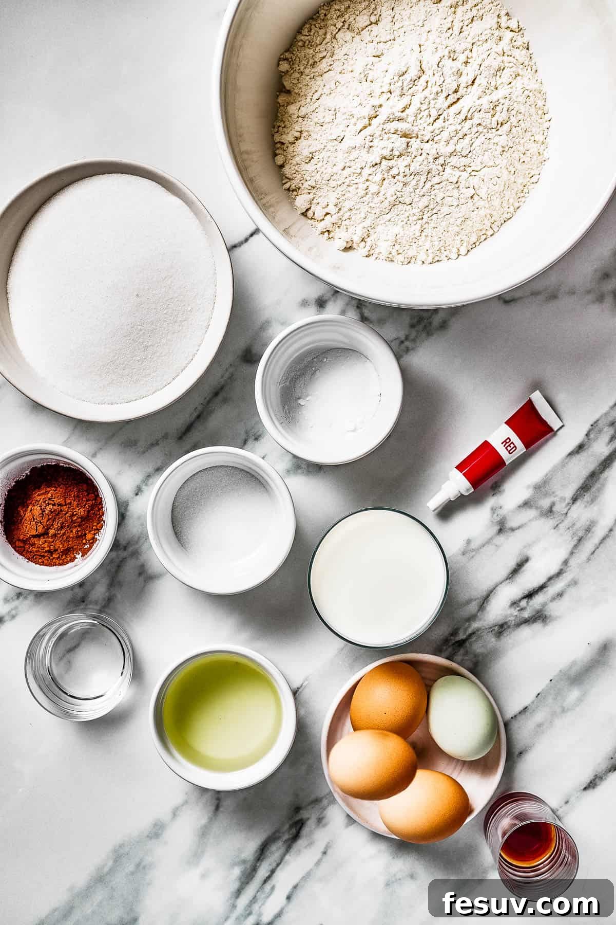 An overhead shot displaying all the dry and wet ingredients laid out on a surface: sugar, baking soda, flour, cocoa powder, salt, milk, red food coloring, vinegar, oil, eggs, and vanilla.
