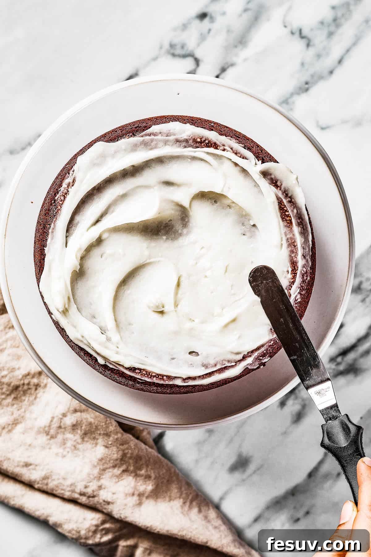 An overhead shot showing the process of frosting a multi-layered red velvet cake with an offset spatula, creating smooth, even layers.
