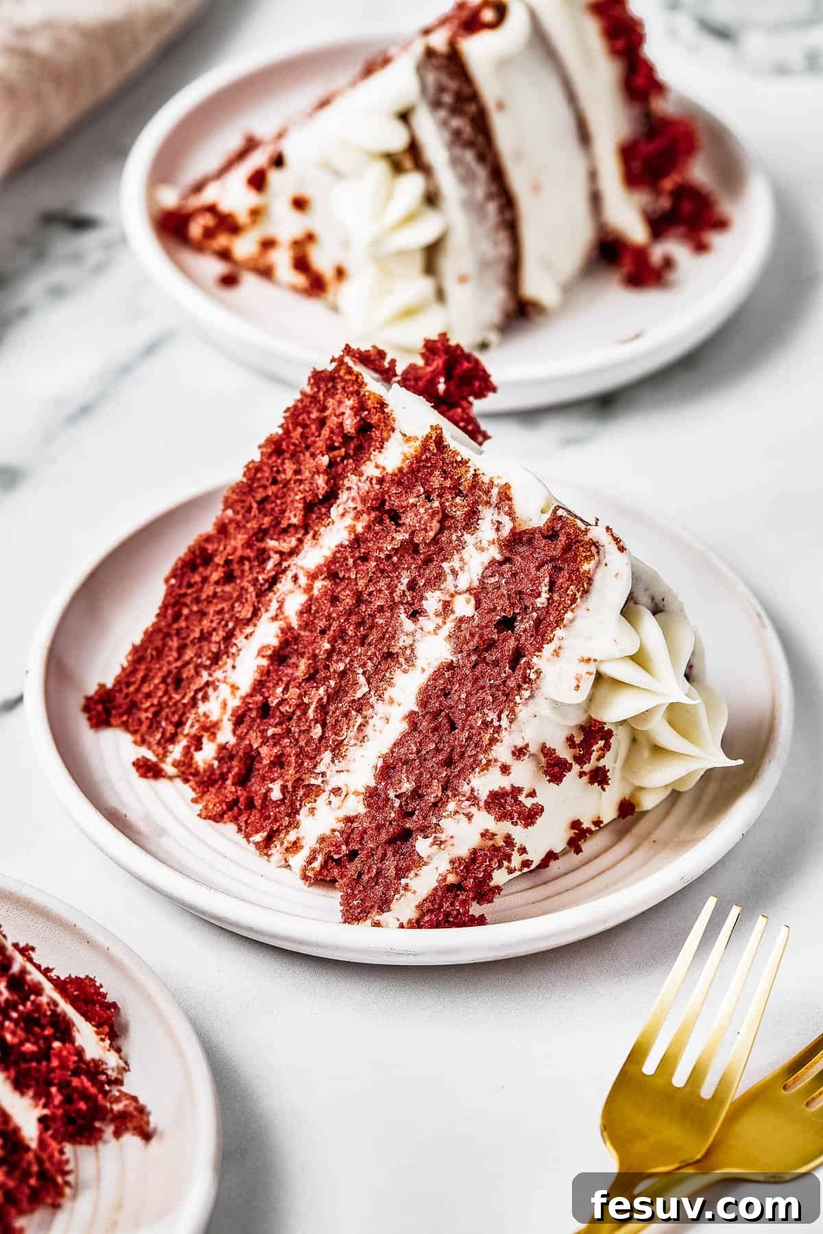 A perfectly portioned slice of red velvet layer cake on a small white plate, accompanied by golden forks on a table.