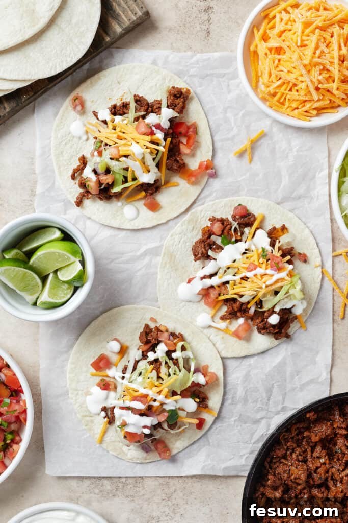 Tortillas on a work surface. Each tortilla is topped with taco meat and various toppings. A bowl of lime wedges and a small plate of cheese are also on the work surface.