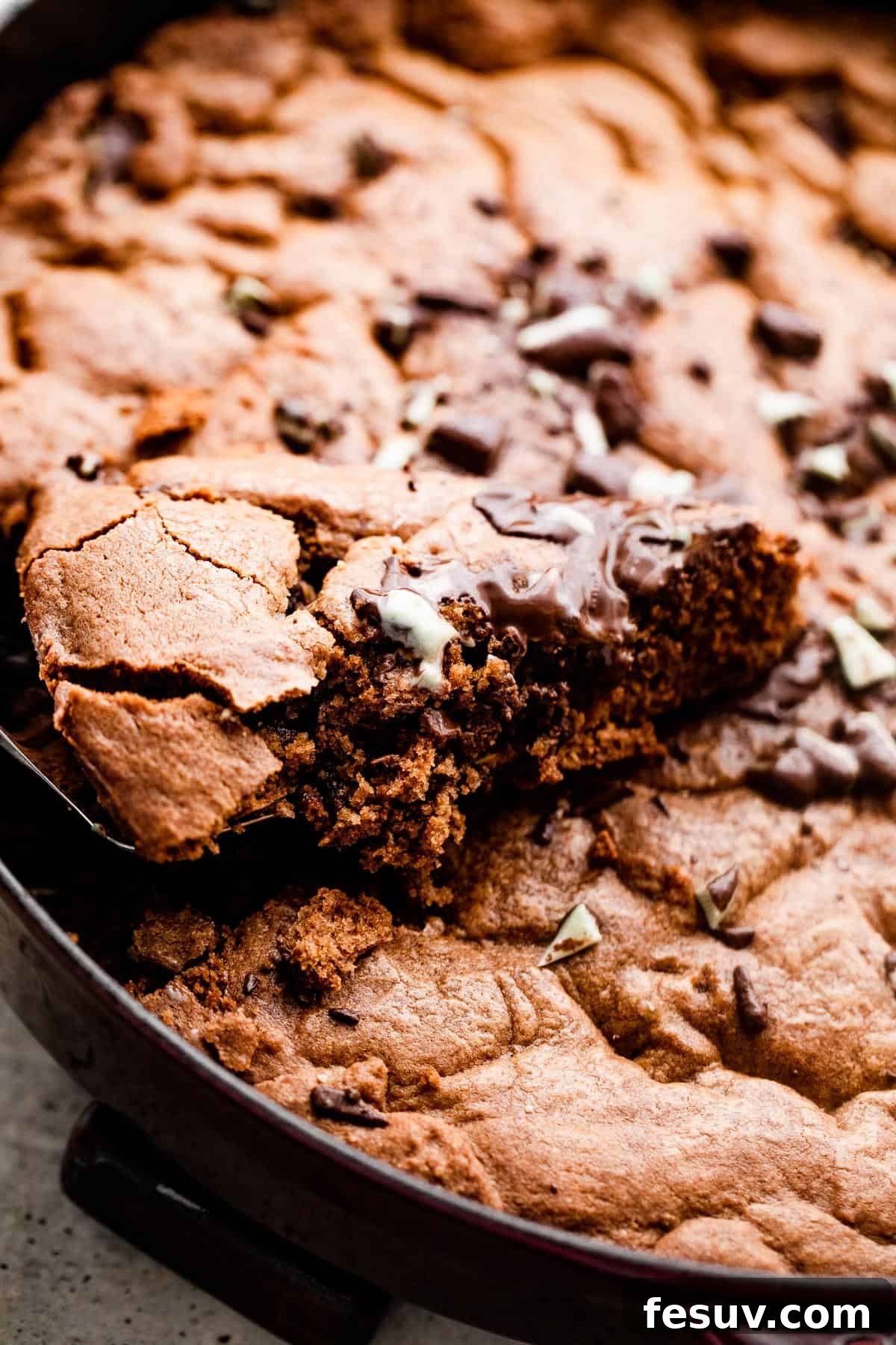 A hand gently pulling out a warm wedge of mint chocolate skillet cookie from a cast iron skillet, showcasing its soft, gooey interior.
