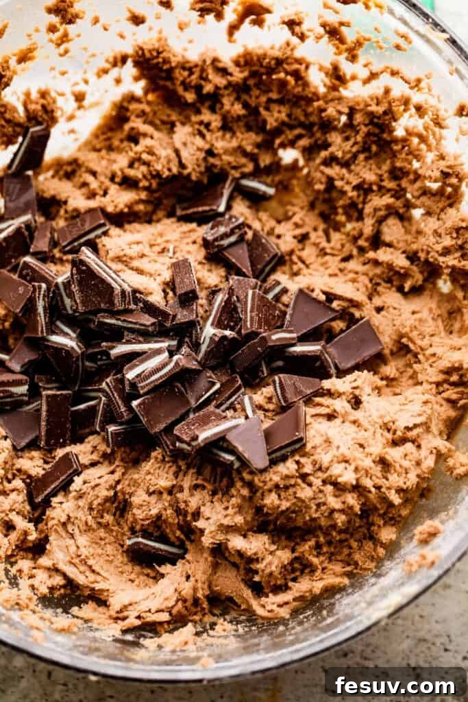 Overhead shot of rich cookie dough in a mixing bowl, with vibrant green and chocolate chopped mints carefully folded into the center of the dough.