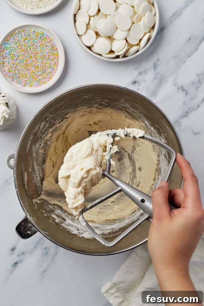 Frosting in a mixing bowl. A woman's hand is lifting a stand mixer attachment covered with frosting out of the bowl.