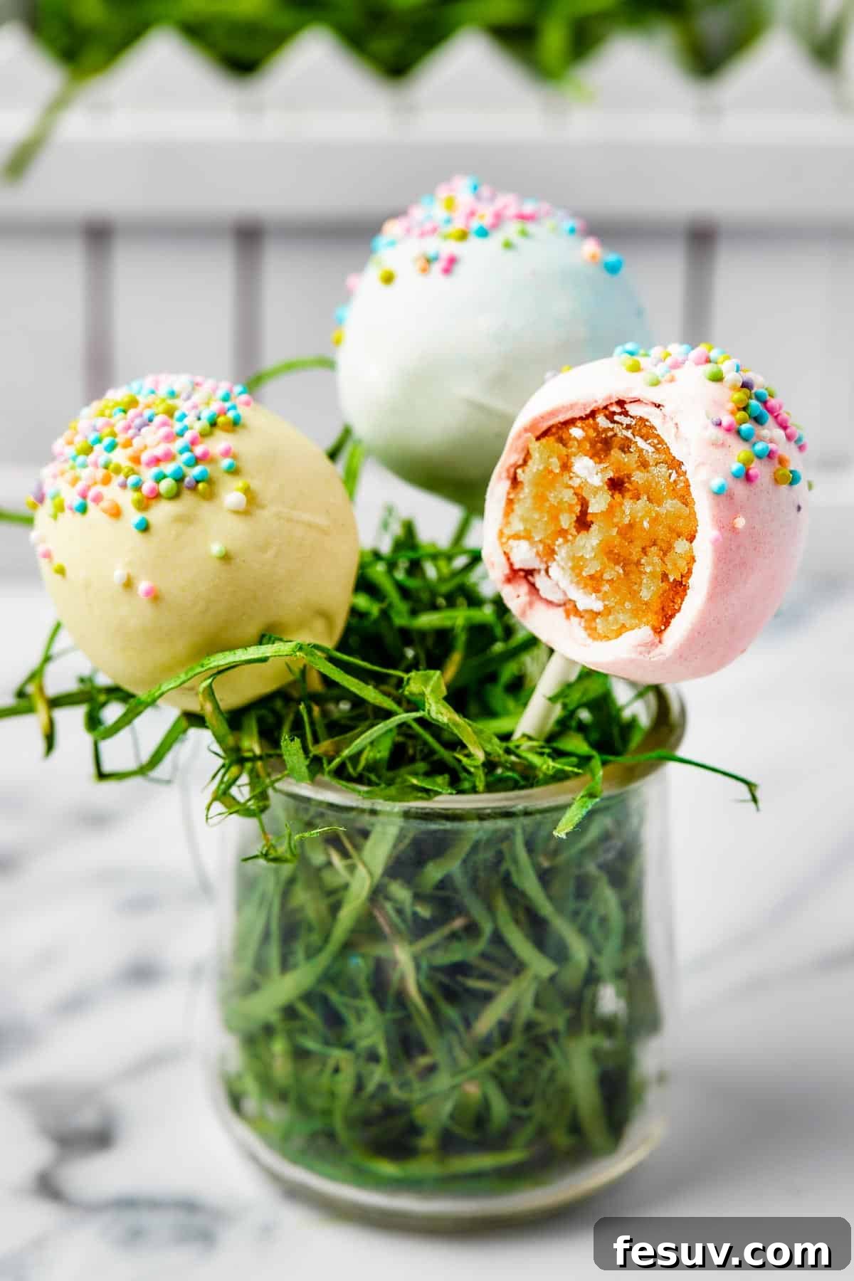 Three cake lollipops in a small glass filled with Easter grass. One of the lollipops has had a bite taken out of it.