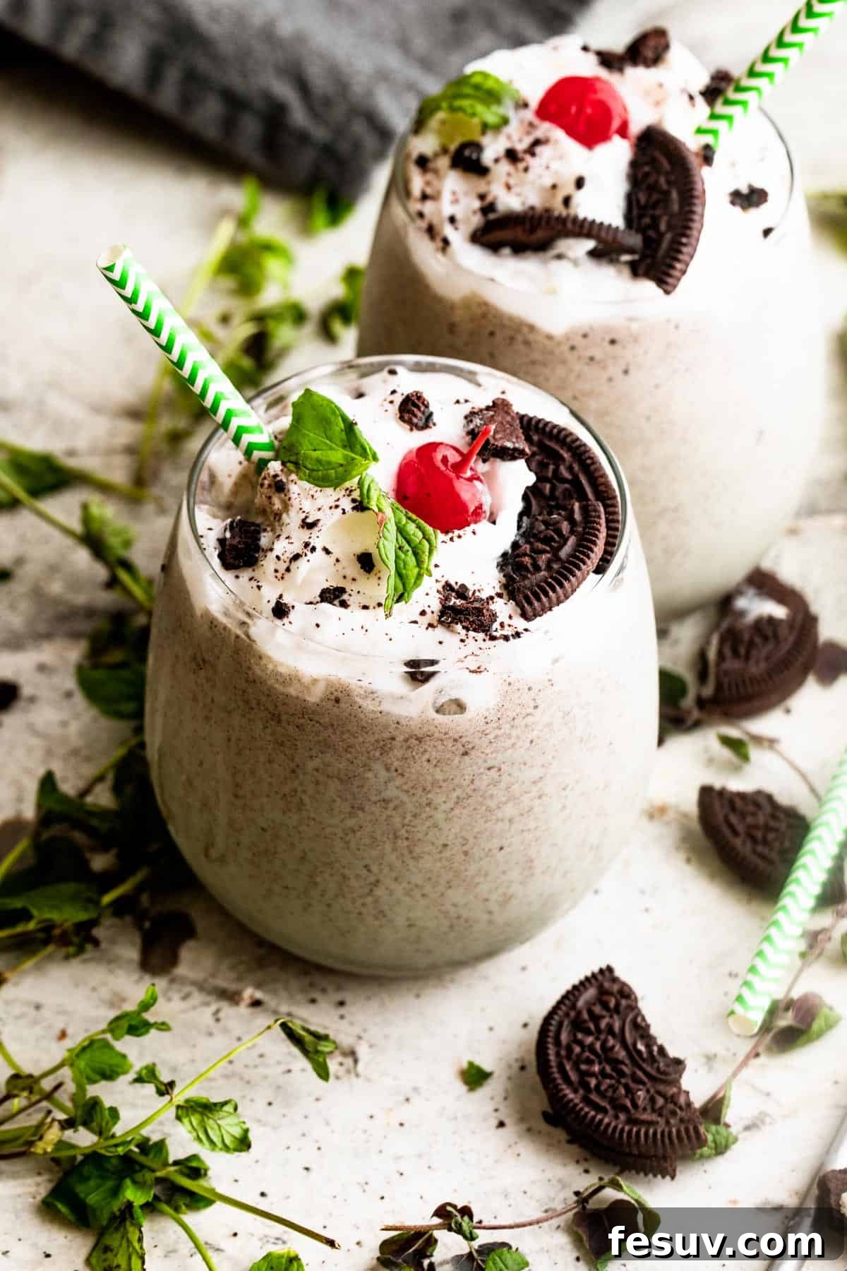 Two clear drinking glasses filled with a thick, green Shamrock Oreo McFlurry, generously topped with fluffy whipped cream, bright red maraschino cherries, and fresh green mint leaves, ready to be served.