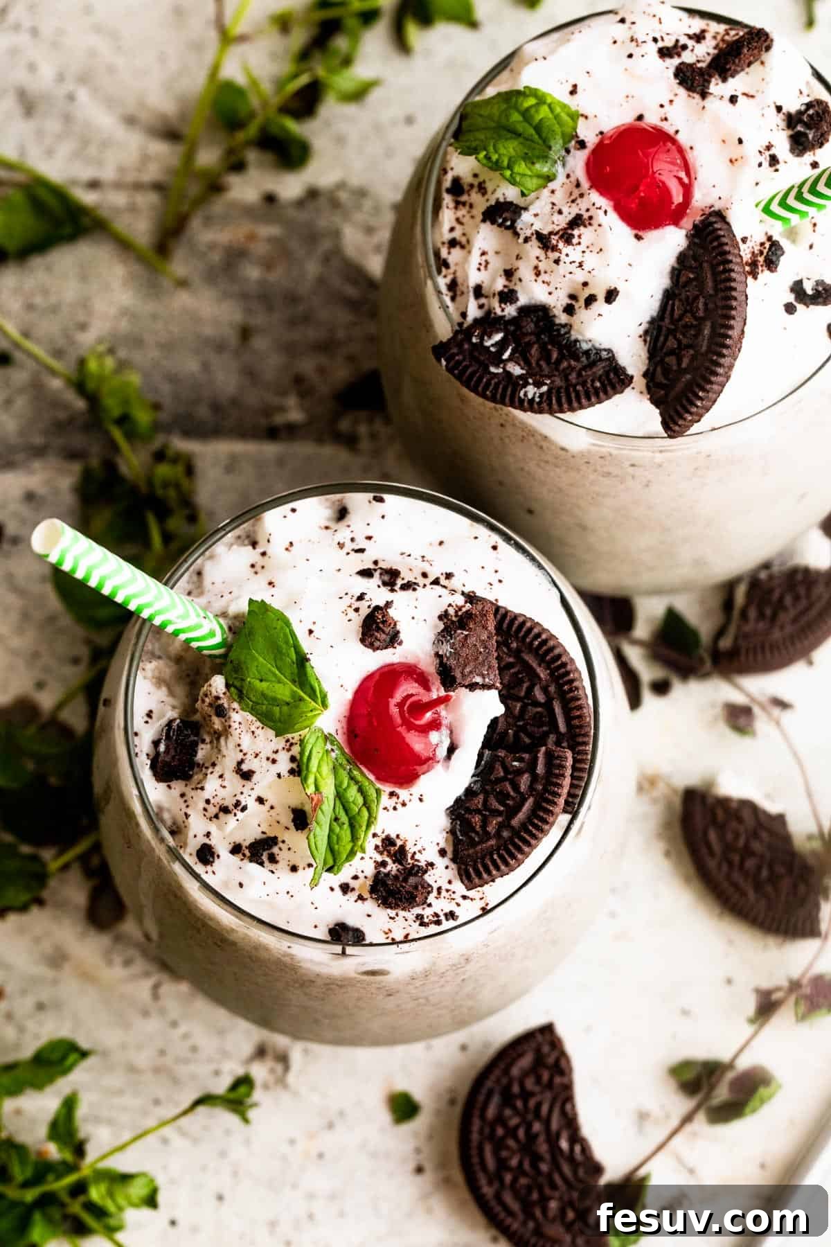 Two drinking glasses filled with a homemade Shamrock Oreo McFlurry, topped with a generous swirl of whipped cream, vibrant maraschino cherries, and fresh mint leaves, ready to be enjoyed.