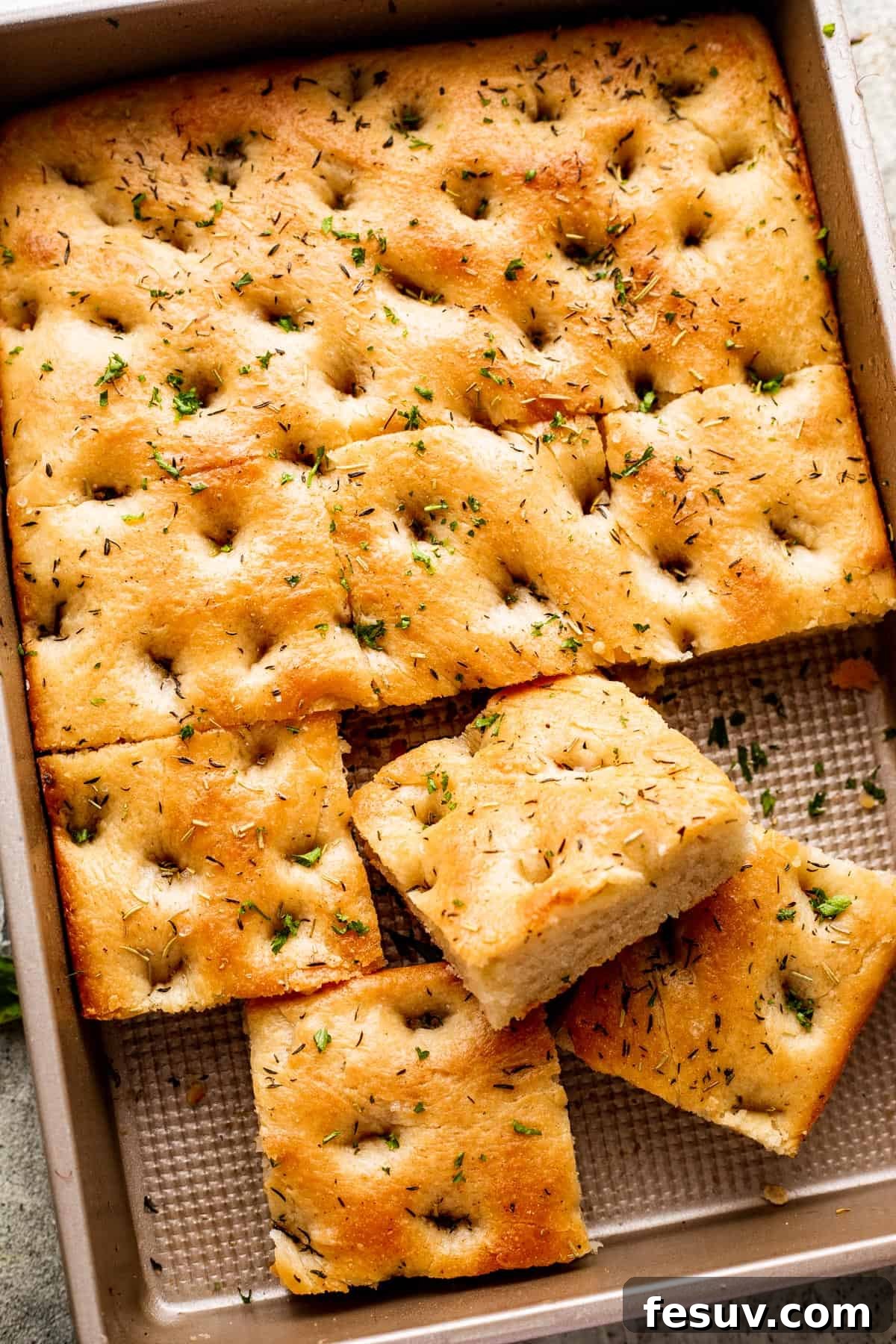 Pillowy Herb-Kissed Garlic Focaccia 2 A large pan-sized loaf of focaccia, with four squares of bread cut away, revealing its soft, airy texture and savory toppings.