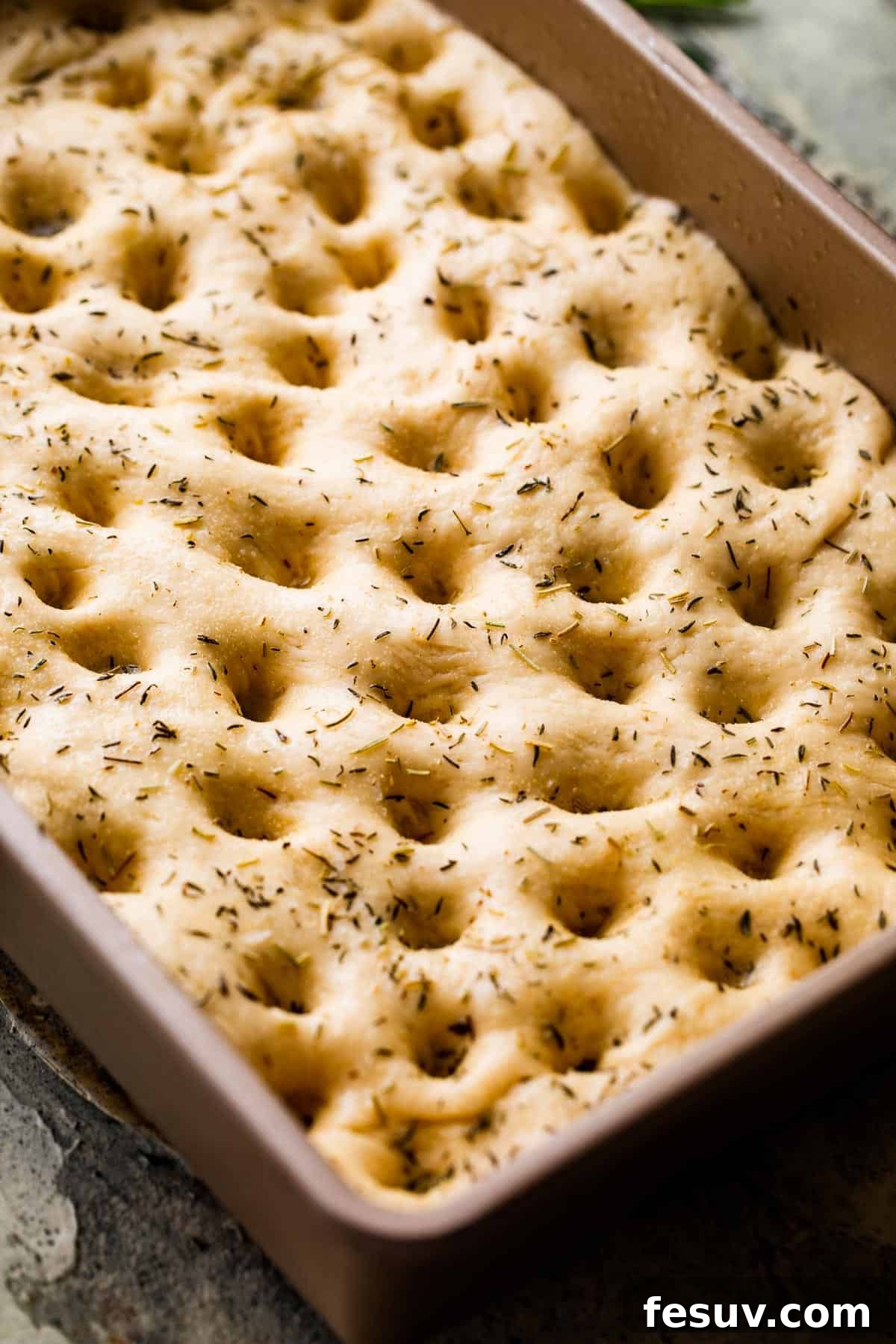 Pillowy Herb-Kissed Garlic Focaccia 7 A pan of unbaked focaccia dough, beautifully poked all over with deep holes and generously sprinkled with fresh herbs and coarse salt.