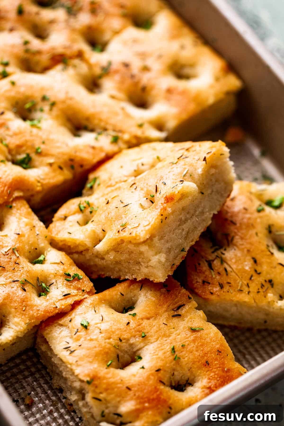 Pillowy Herb-Kissed Garlic Focaccia 9 A large pan-sized loaf of freshly baked focaccia, with four square pieces perfectly cut away and presented on a wooden board.