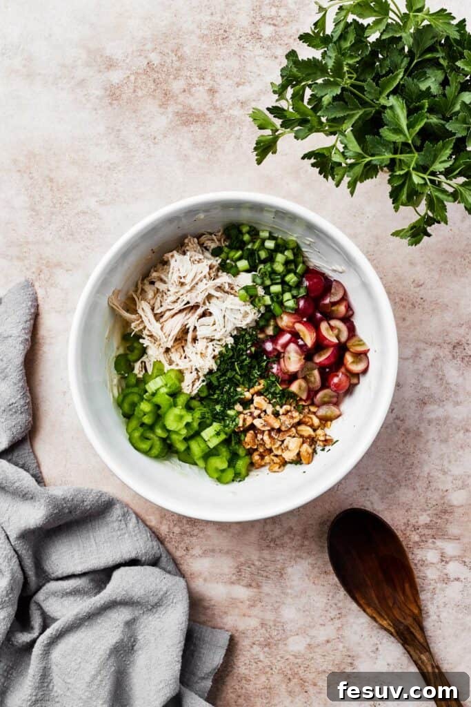 A white mixing bowl with shredded chicken, vegetables, grapes, and other ingredients.