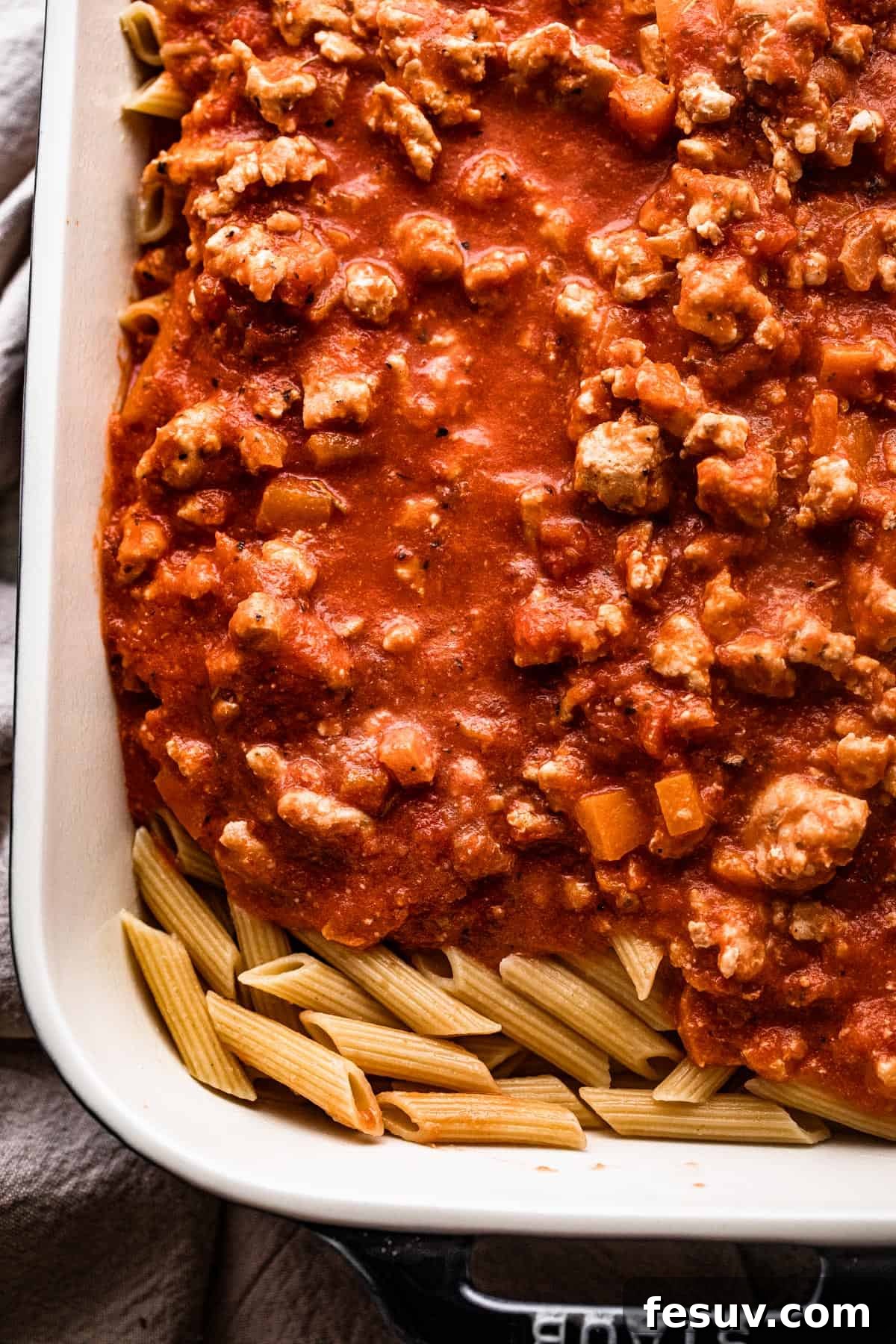 Velvet Penne Vodka Bake 5 A generous serving of ground turkey tomato sauce being poured over a bed of al dente penne pasta in a large mixing bowl, ready to be combined and baked into a comforting casserole.