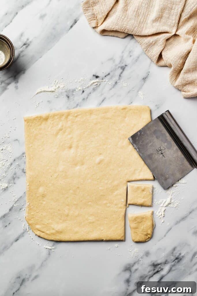 French Quarter Powdery Delights 7 A square of rolled-out yeast dough on a marble work surface, with small square pieces cut out of it. A bench scraper is resting on the dough.