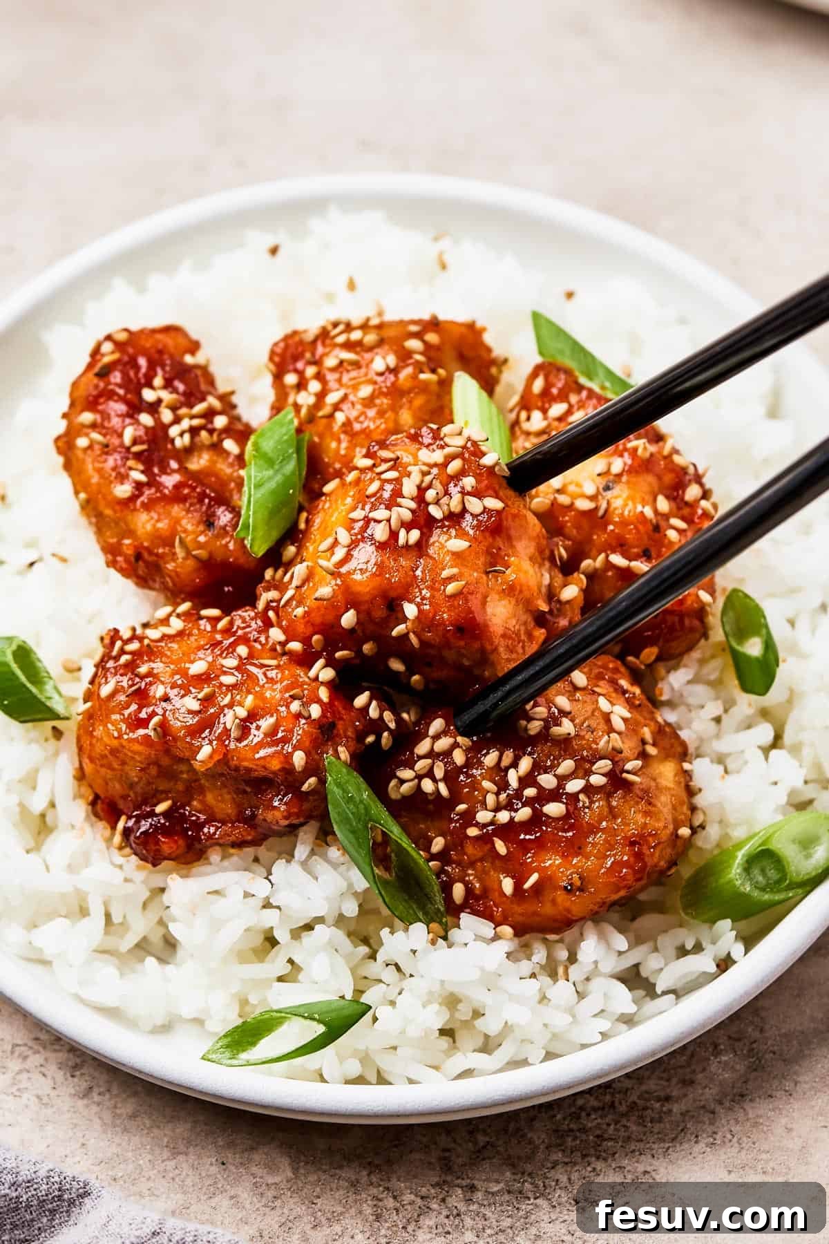 A small plate of takeout-style sweet and sour chicken over fluffy white rice, with chopsticks holding one piece of chicken, highlighting its crispy texture and glistening sauce.