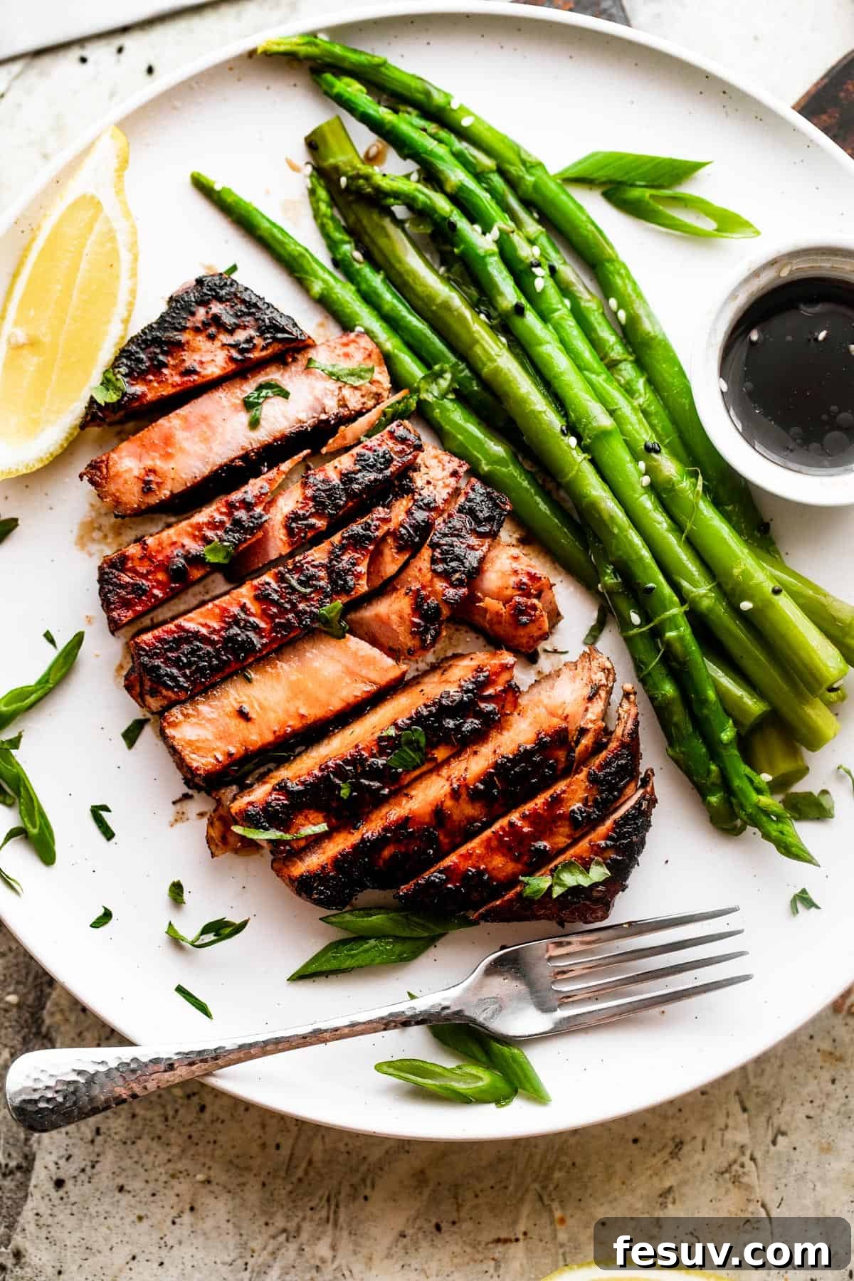 Overhead shot of a sliced blackened balsamic tuna steak served on a white dinner plate with bright green asparagus stalks, a small dipping sauce, and fresh lemon slices.