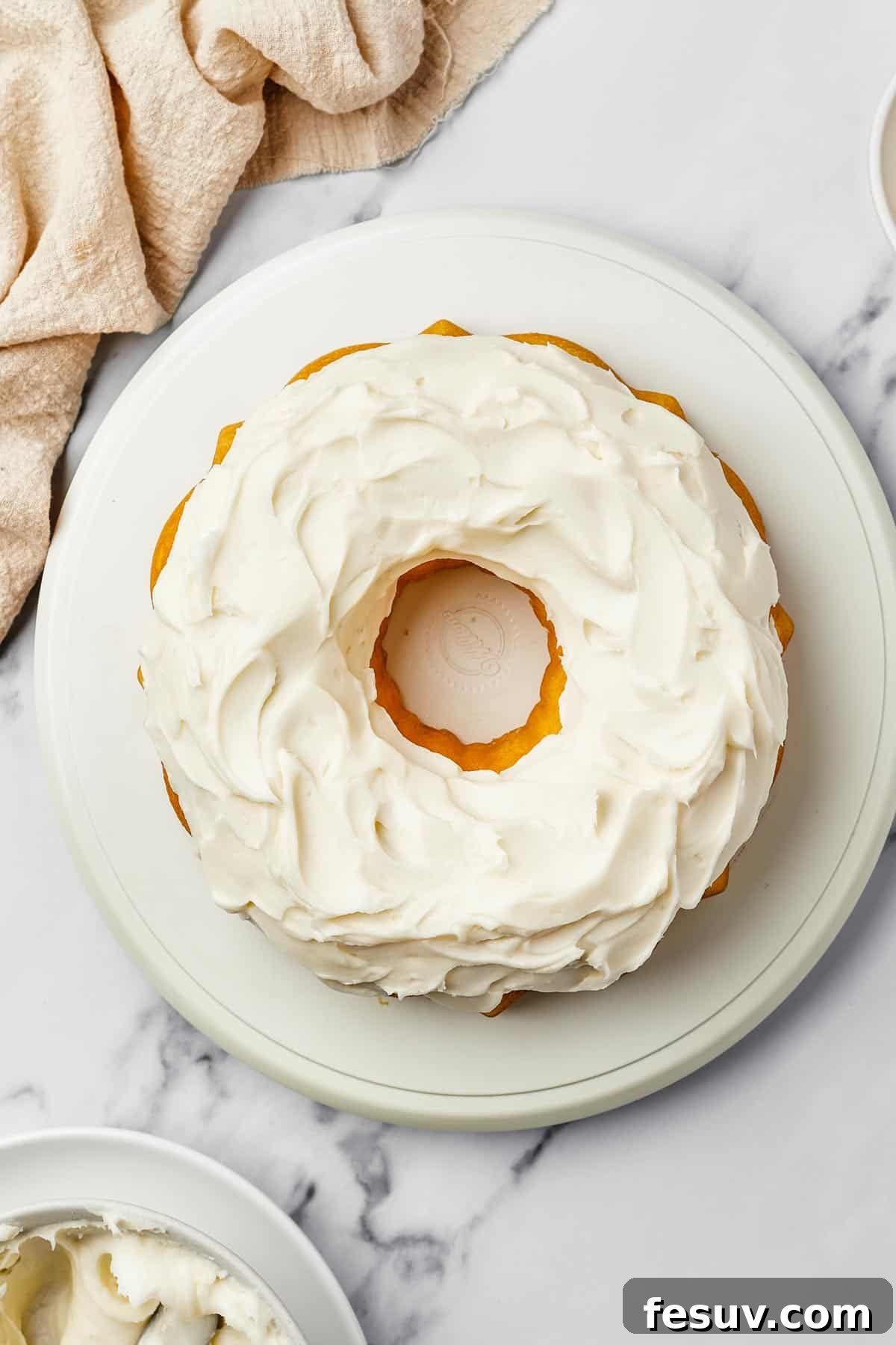Overhead shot of a bundt cake iced with thick, creamy frosting. A bowl of frosting and dish of lemon zest are nearby on the table.