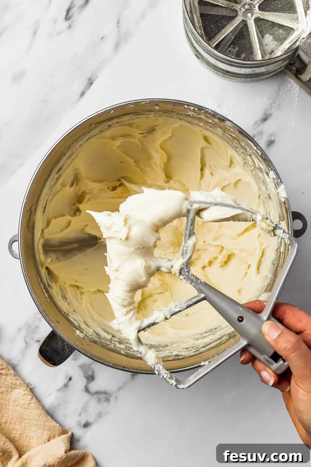 A metal mixing bowl with whipped frosting inside. A woman's hand is lifting a beater attachment out of the frosting.