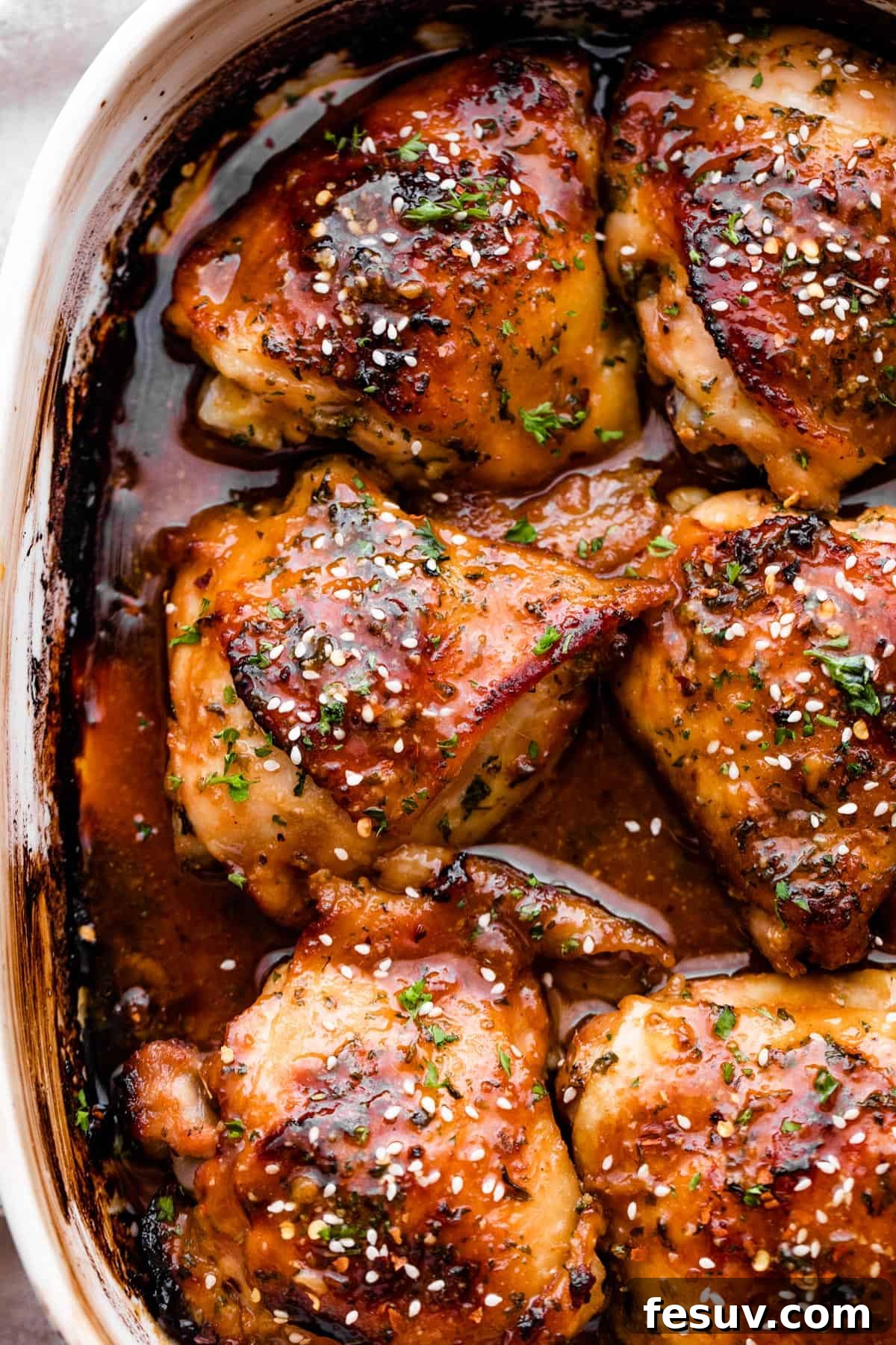 Overhead shot of a baking dish filled with six beautifully golden-brown honey garlic baked chicken thighs, generously garnished with toasted sesame seeds and fresh parsley.
