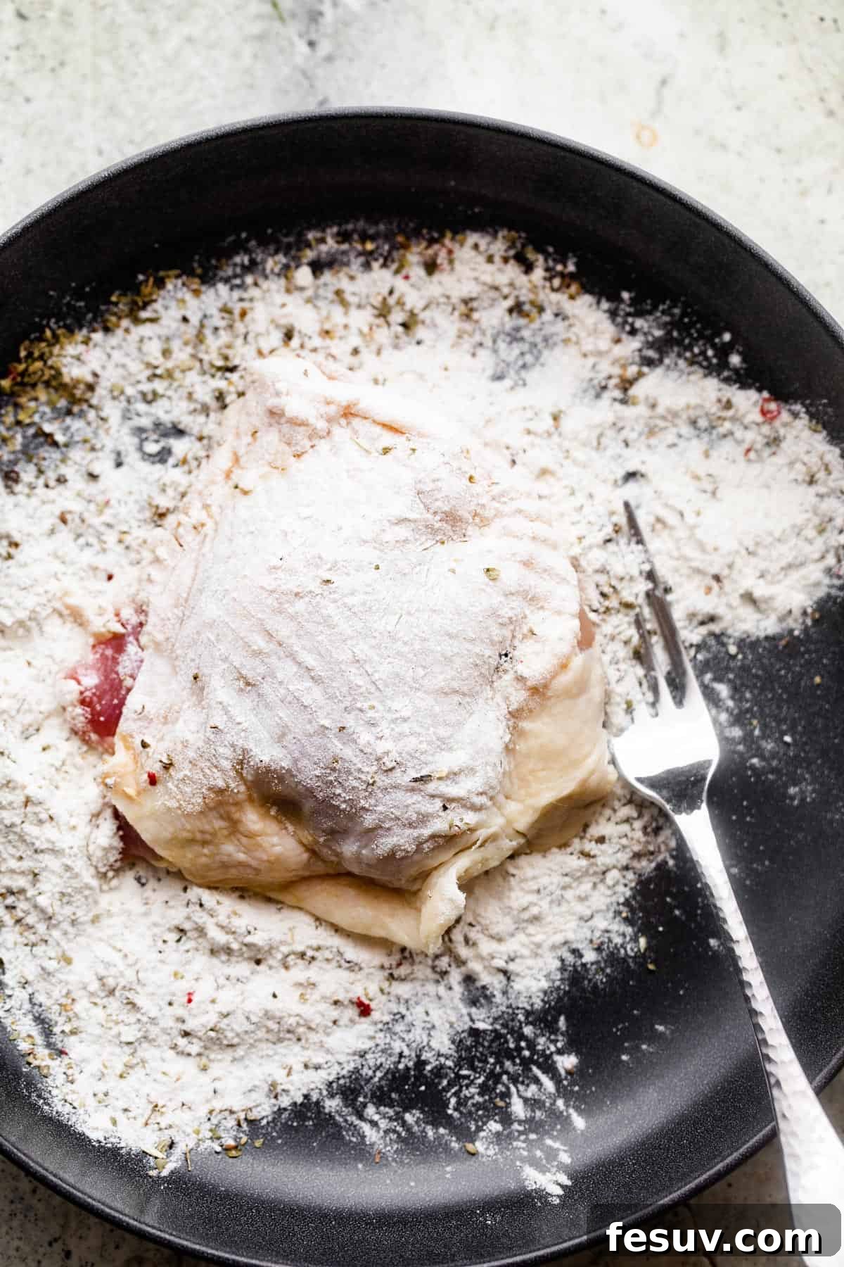 Raw bone-in, skin-on chicken thigh placed on a black plate with a mound of seasoned flour next to it, indicating the dredging process.