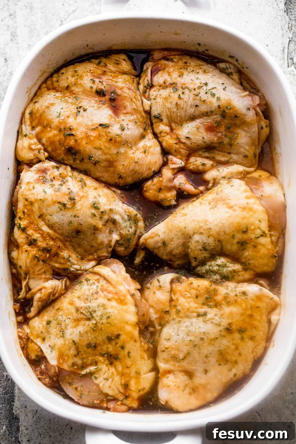 Overhead image of a white baking dish containing six raw, flour-dredged bone-in, skin-on chicken thighs, neatly arranged and ready for the sauce and baking.