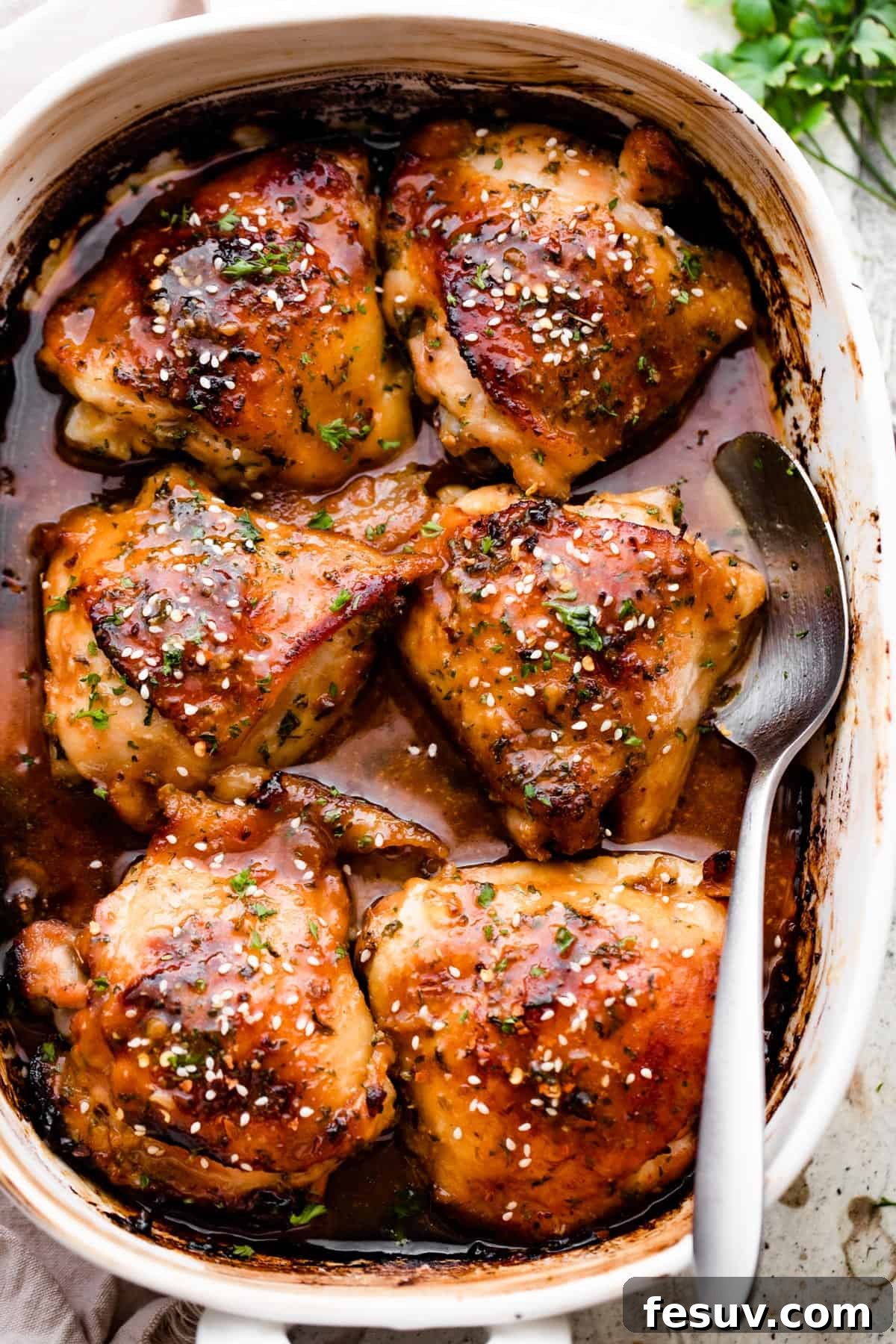 Overhead shot of a baking dish with six perfectly honey-garlic baked chicken thighs, beautifully golden-brown, glistening with sticky sauce, and generously sprinkled with toasted sesame seeds.