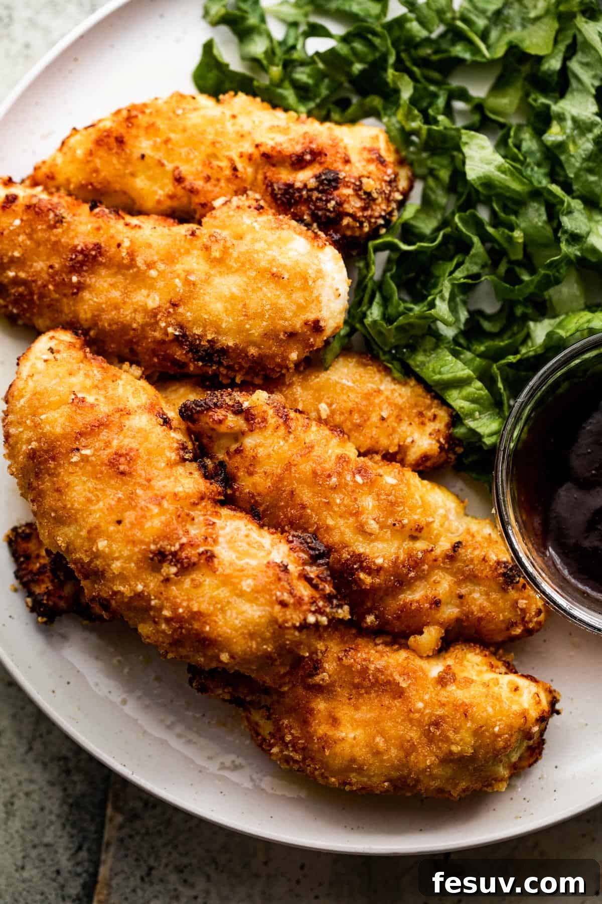 Restaurant Quality Crispy Air Fryer Chicken 3 A close-up overhead shot of a dinner plate featuring crispy air fryer chicken tenders, fresh greens, and a bowl of sweet and savory BBQ sauce.
