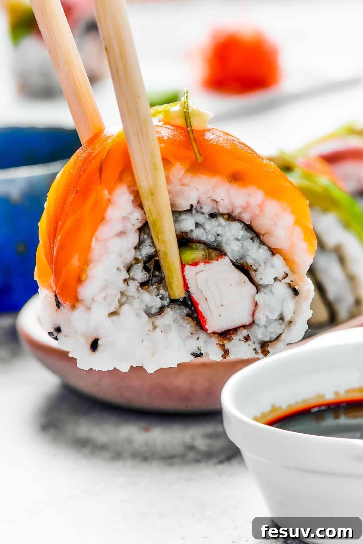 Rainbow Sushi Masterpiece 2 A bite of sushi held in a pair of chopsticks. A small plate of sushi and a container of soy sauce is in the background.