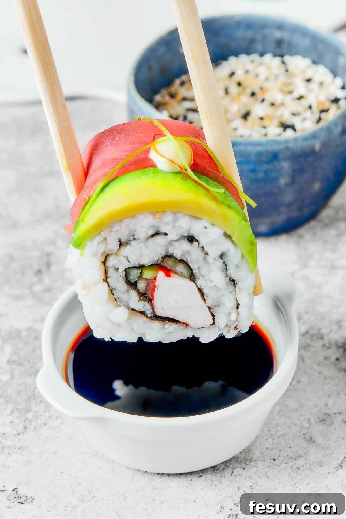 Rainbow Sushi Masterpiece 12 A bite of sushi held in a pair of chopsticks. A small plate of sushi and a container of soy sauce is in the background.