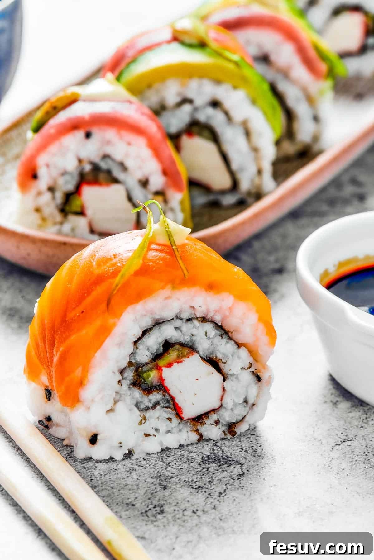 Rainbow Sushi Masterpiece 13 A bite of sushi set on a surface. A small plate of sushi and a container of soy sauce is in the background.