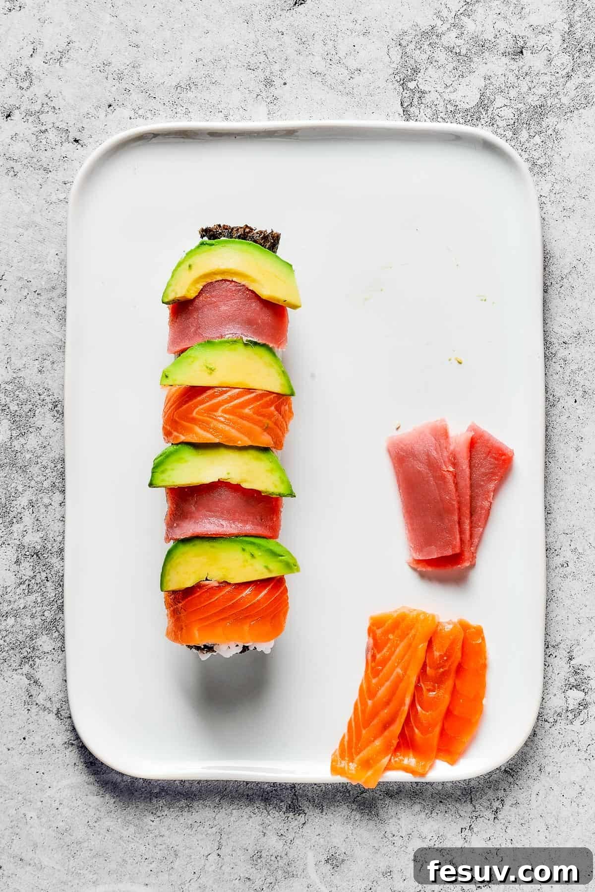 Rainbow Sushi Masterpiece 10 Overhead shot of a roll of sushi, topped with alternating slices of avocado, tuna, and salmon. Extra slices of tuna and salmon are on the platter.