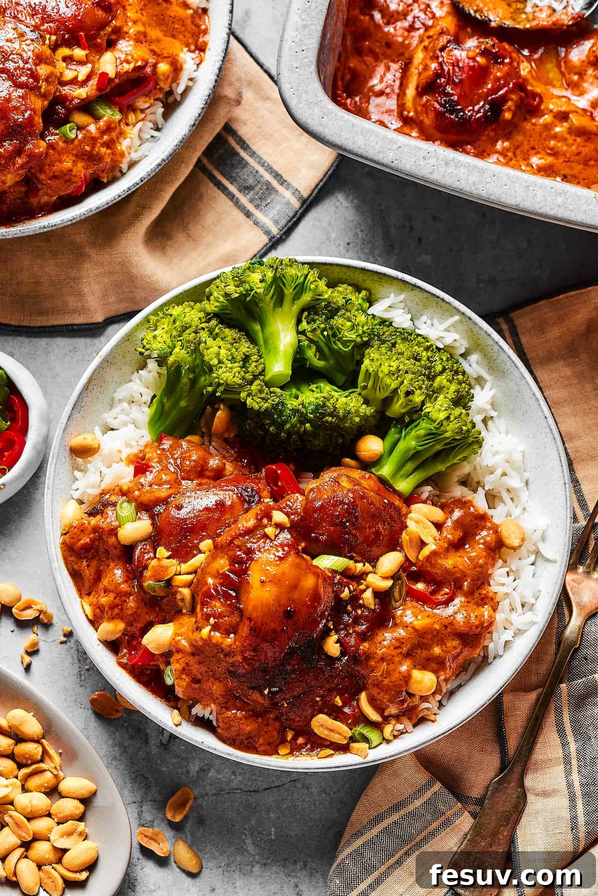 Simple Peanut Butter Chicken with crushed tomatoes, soy sauce, and garlic is a breeze to make. Serve this saucy chicken over rice, noodles, or cauliflower rice, and watch it disappear! Peanut chicken served with broccoli over rice. A baking dish of the chicken is nearby on the table, along with garnishes and more plates.