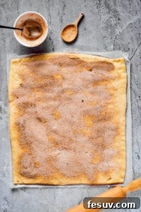 A work surface with a large rectangle of dough, a rolling pin, and a small bowl of cinnamon sugar nearby. Cinnamon sugar has been sprinkled generously all over the dough.