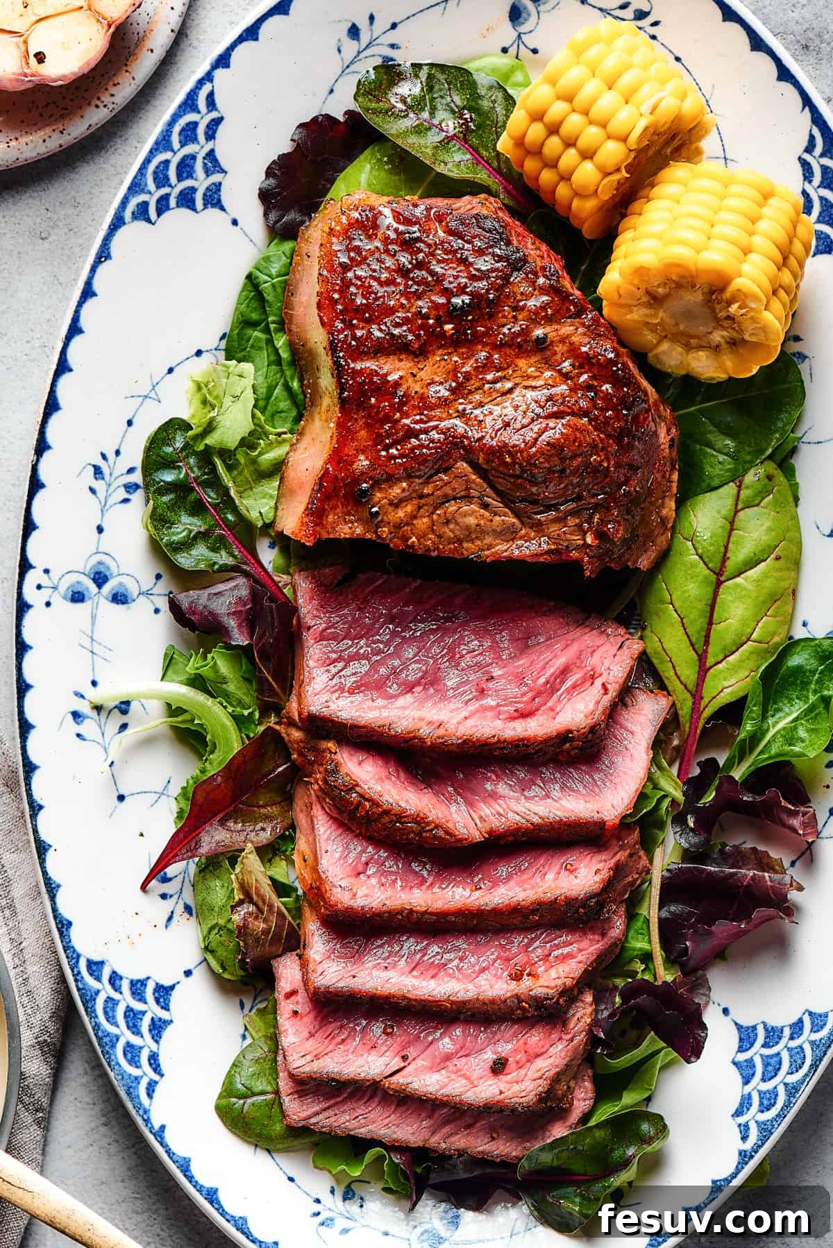 A large blue steak artfully arranged on a bed of fresh greens, garnished with vibrant corn on the cob. Half of the steak is expertly sliced and fanned out, proudly showcasing its stunning, ultra-rare crimson interior.
