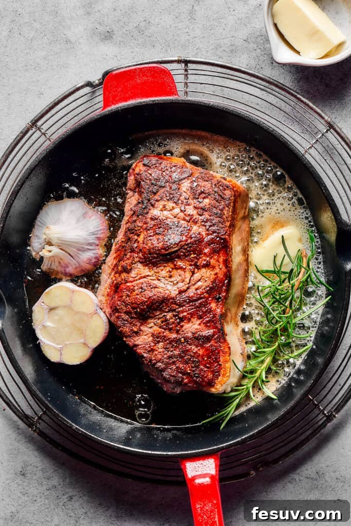 A prime sirloin steak searing beautifully in a hot cast-iron pan, alongside melting butter, a fresh sprig of rosemary, and a halved head of garlic, infusing the cooking oil with rich aromas.