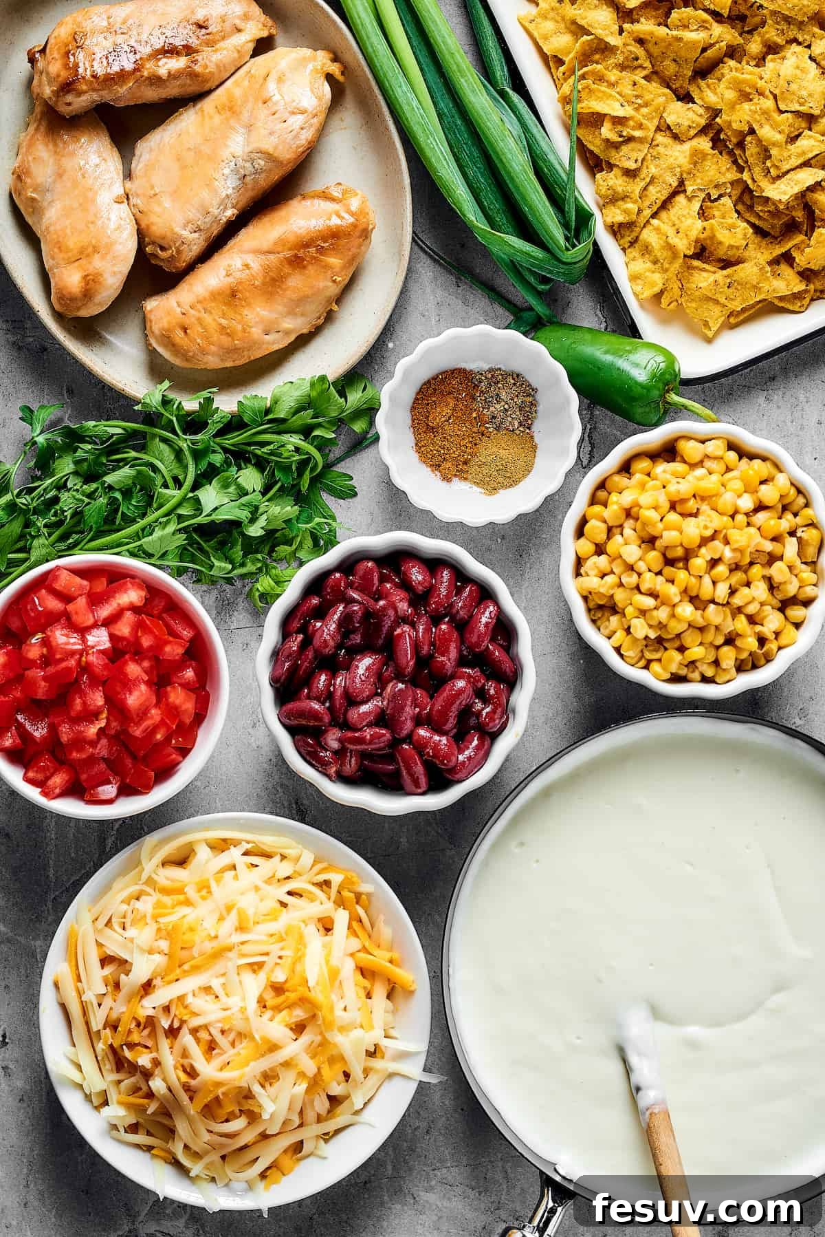 Fiesta Chicken Casserole 3 Overhead view of various ingredients laid out: cooked chicken, green onions, crushed chips, spices, chopped tomatoes, beans, corn, two types of cheese, and white sauce.