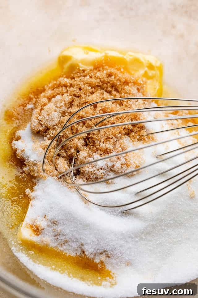 Mixing together softened butter, both brown and granulated sugars, and vanilla extract in a bowl, showing the creamy texture of the cookie dough base.