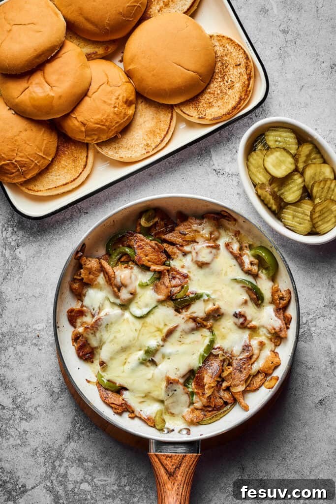 A tray of sandwich buns and a small dish of sliced dill pickles next to a skillet with cooked steak, onion, and bell pepper, topped with melted cheese.