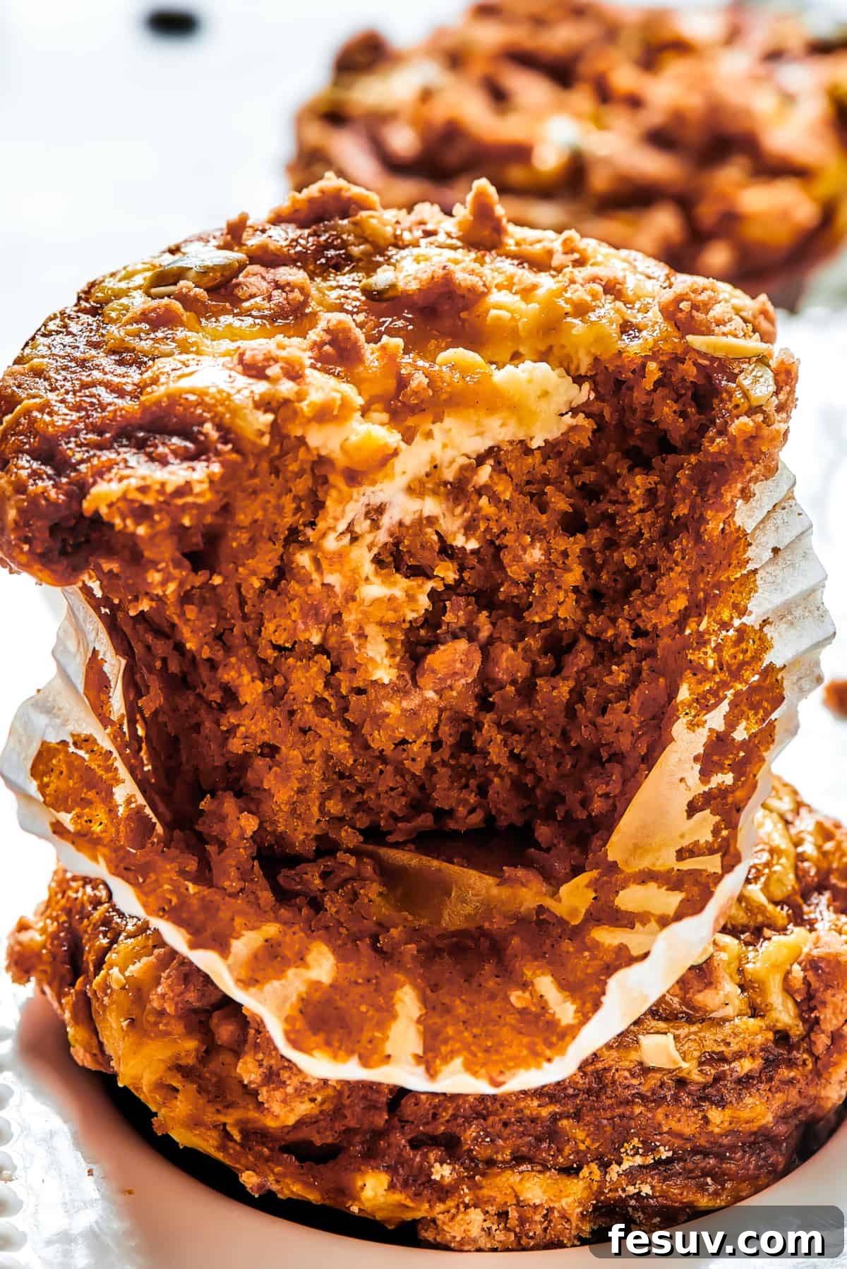 Pumpkin Cream Cheese Muffins are a heavenly mix of spiced pumpkin cake and smooth cream cheese swirl, with a buttery, streusel-like topping. A pumpkin muffin broken in half with the paper muffin cup peeled away.