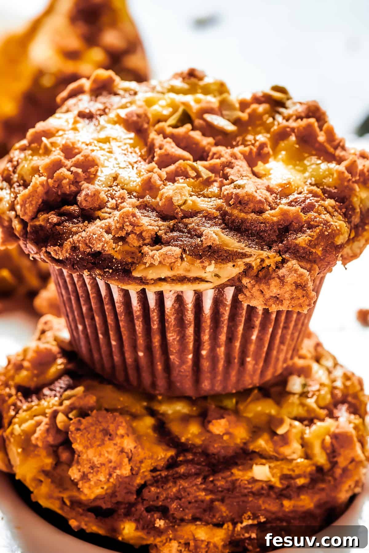 Pumpkin Cream Cheese Muffins are a heavenly mix of spiced pumpkin cake and smooth cream cheese swirl, with a buttery, streusel-like topping. A pumpkin cream cheese muffin set atop of another muffin, and arranged on a work surface.