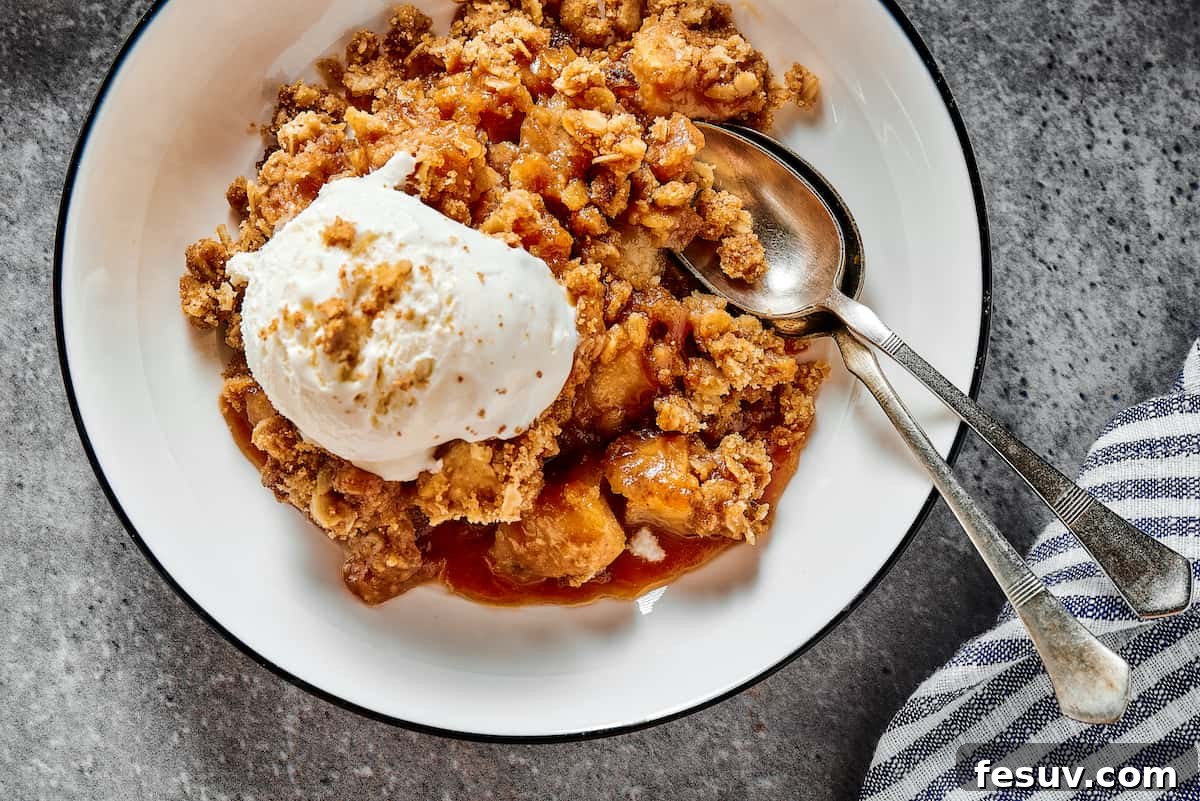 A serving of homemade apple crisp topped with a scoop of melting ice cream and a drizzle of caramel, presented in a small white bowl.