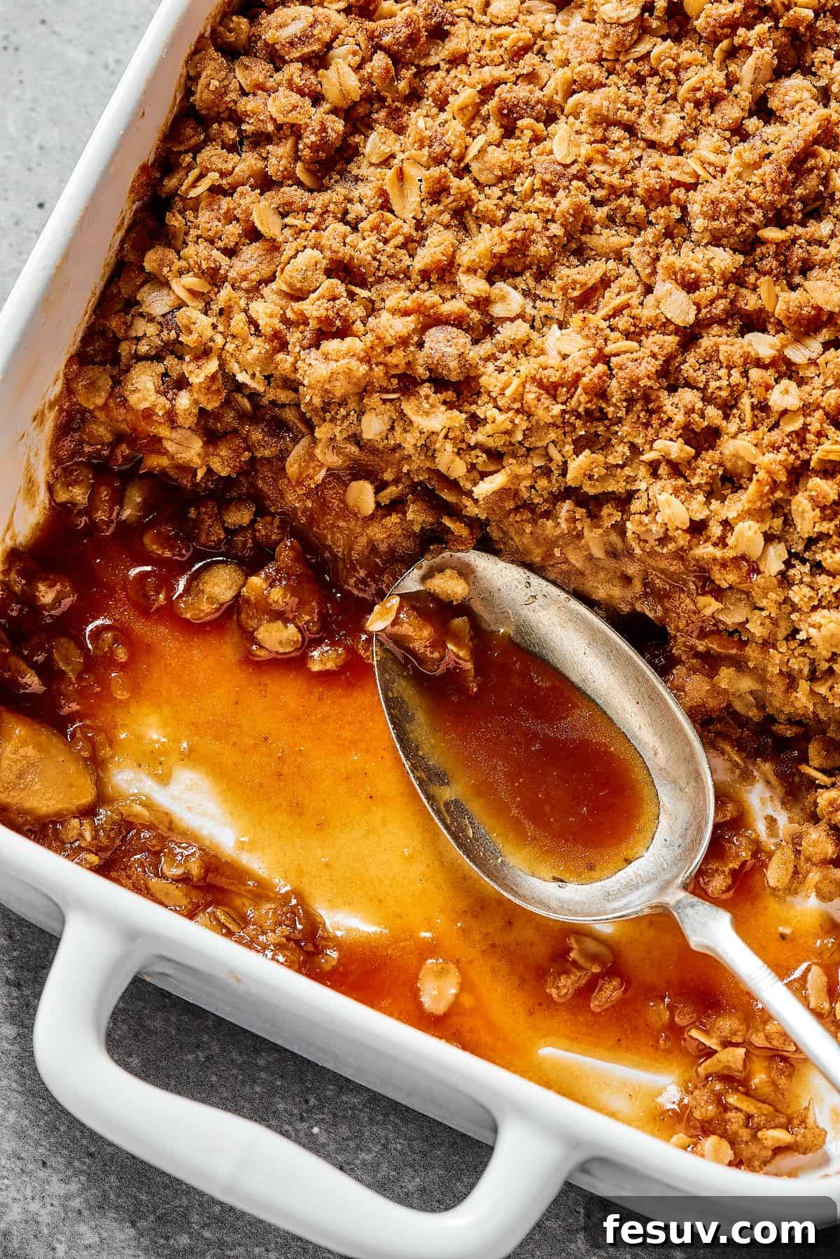 A close-up of a partially eaten apple crisp in a white casserole dish, showcasing the bubbling spiced apple filling beneath the golden, crumbly oat topping. A spoon rests beside the dish.