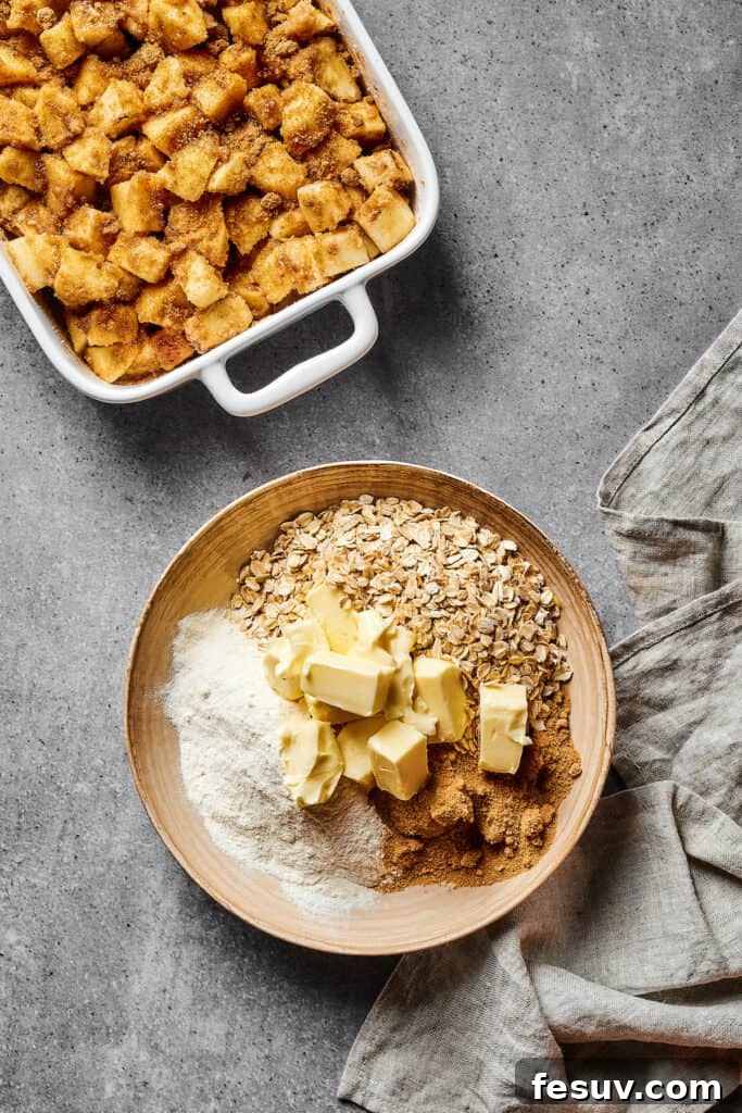 Ingredients for the crisp topping – rolled oats, cubed cold butter, flour, and brown sugar – are arranged next to a bowl of spiced apples in a casserole dish.